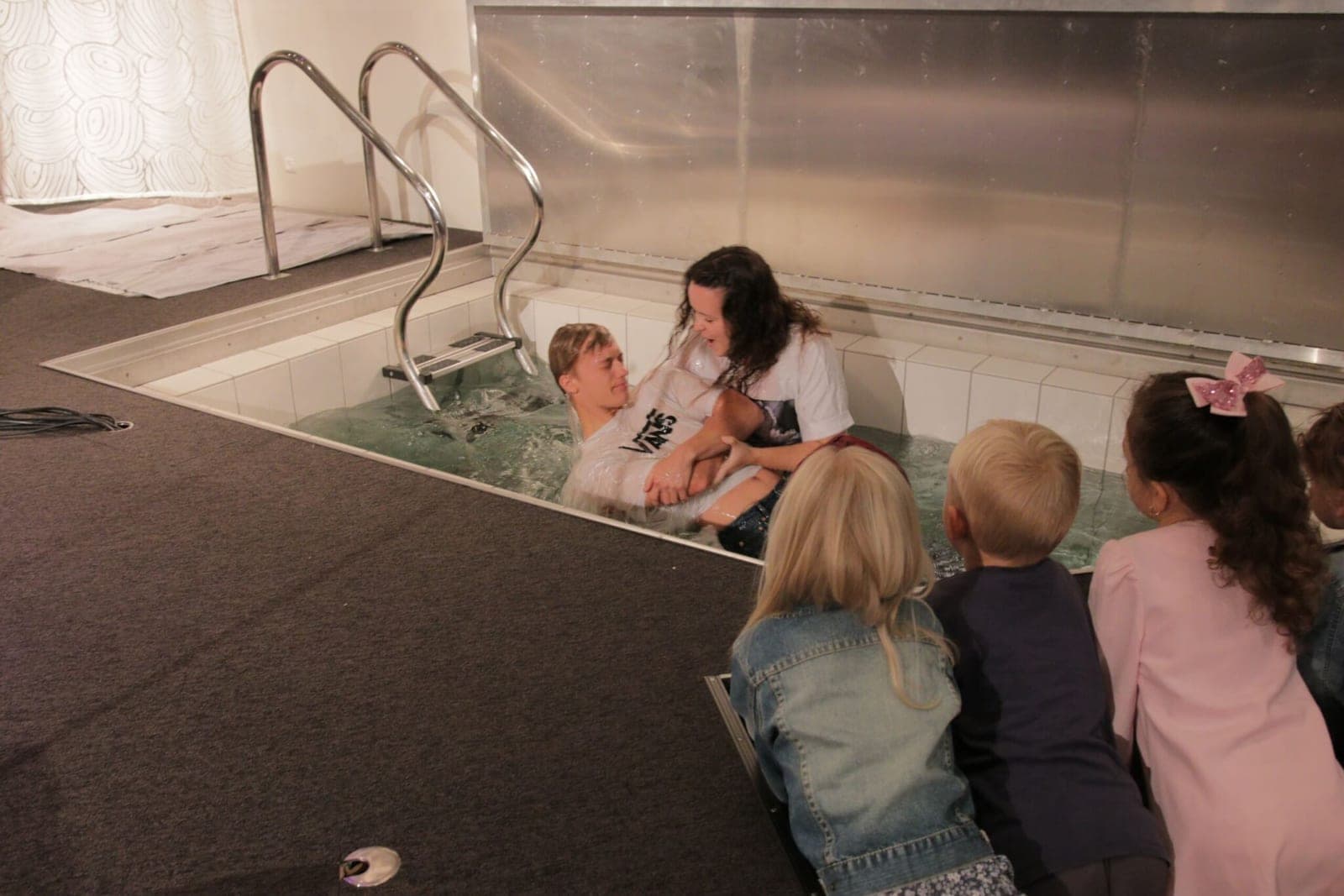 A young person being baptised by full immersion in the church baptismal pool while children watch